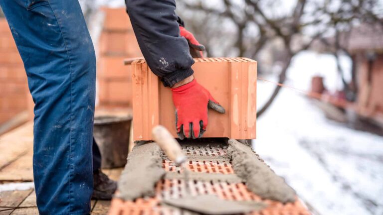 A builder with gloved hands constructs a brick wall during the winter